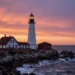 A dramatic seascape photograph of the Portland Head Lighthouse in Maine, USA, at sunset. The white stone lighthouse stands on a rocky cliff overlooking the Atlantic Ocean. Waves crash against the rocks below, and the sky is painted in shades of orange, pink and purple. The scene is majestic, with a sense of solitude and maritime history. High resolution, hyper-realistic, cinematic lighting.