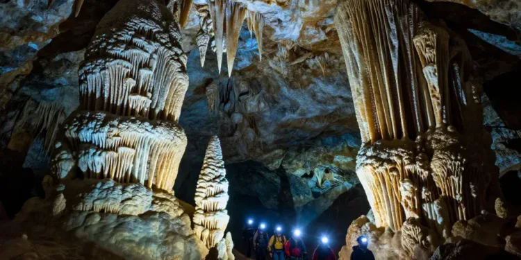 A dramatic, photorealistic image of an underground cave in North America, massive stalactites and stalagmites, crystal formations glowing with soft blue and amber light, a small group of explorers with headlamps walking on a narrow path, deep darkness surrounding, high detail, cinematic lighting, 8K resolution.