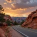 A panoramic view of a red rock canyon at sunset, with a winding road leading into the distance, pine trees on the left, and a dramatic sky with clouds, evoking the spirit of an American national park road trip, photorealistic style, 16:9 aspect ratio.