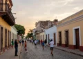A panoramic view of a historic North American city street, blending colonial architecture with modern elements, cobblestone path, colorful buildings with wrought-iron balconies, a street musician playing a trumpet, and a diverse crowd walking, under a warm sunset sky, photorealistic style.