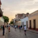 A panoramic view of a historic North American city street, blending colonial architecture with modern elements, cobblestone path, colorful buildings with wrought-iron balconies, a street musician playing a trumpet, and a diverse crowd walking, under a warm sunset sky, photorealistic style.