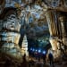 A dramatic, photorealistic image of an underground cave in North America, massive stalactites and stalagmites, crystal formations glowing with soft blue and amber light, a small group of explorers with headlamps walking on a narrow path, deep darkness surrounding, high detail, cinematic lighting, 8K resolution.